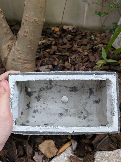 屋外の地面を背景に、焼けたような黒い丸太を模した長方形セメント製プランターを上から覗き込んだ様子。プランターの内側の質感と、底にある水抜き穴がはっきりと見える。プランターは手に持たれている。